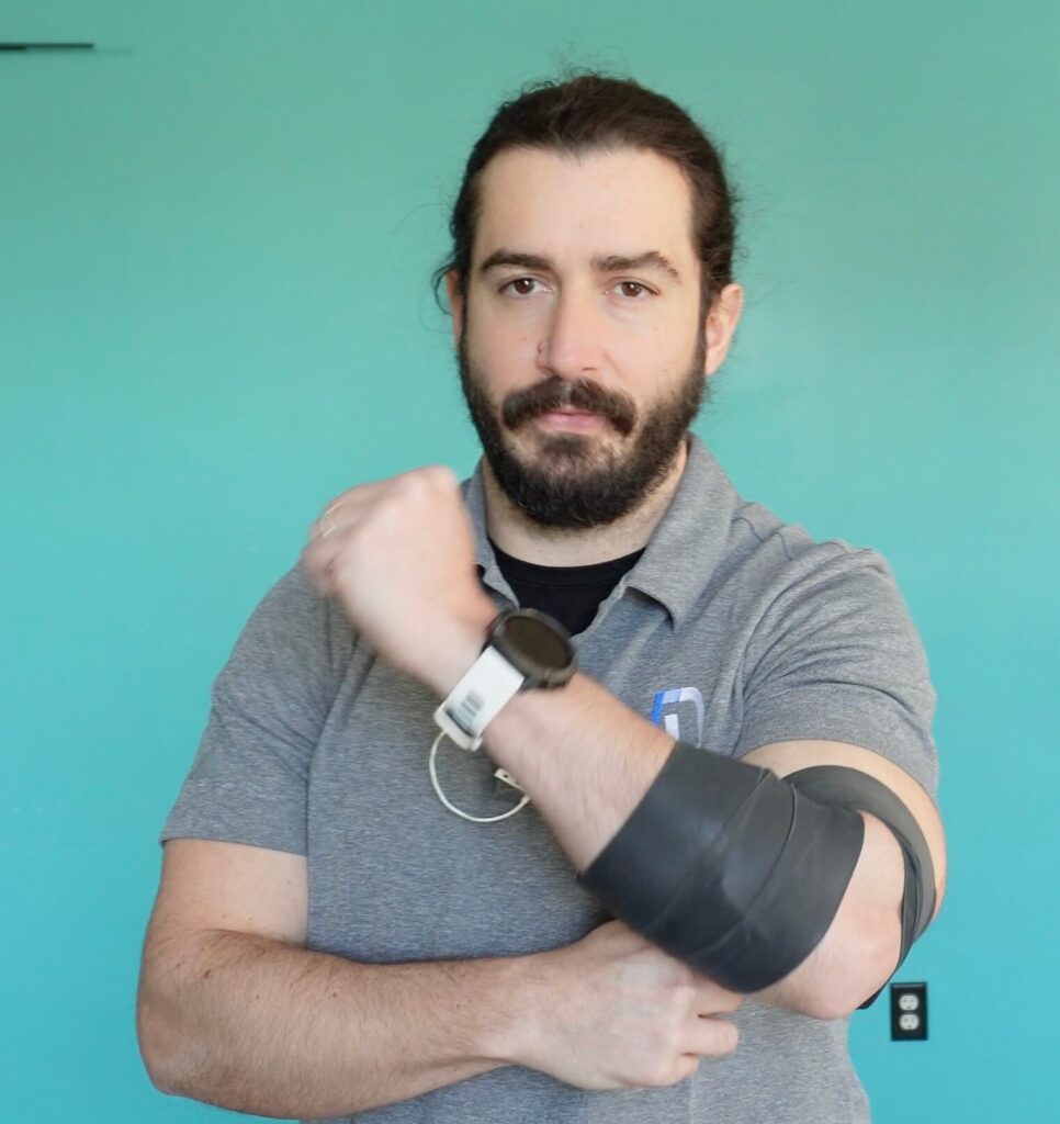 Dr. John with muscle floss tightly wrapped around his left elbow bent across his body as he flexes